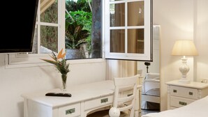 Quarto com escrivaninha, cadeira, televisão, mesa de cabeceira com luminária e janela com cortinas.