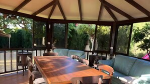 Varanda coberta com mesa e cadeiras de madeira, um sofá azul claro e vista para o verde.