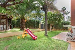 A playground with a red slide, yellow and blue play structures, and palm trees.
