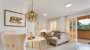 Uma sala de estar com sofá branco, mesa de jantar com uma cadeira, ilustração emoldurada de um animal e luminária pendente.