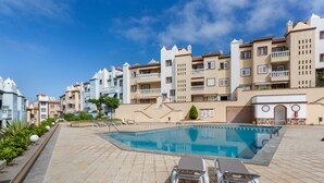 Piscina com espreguiçadeiras, um deck revestido de azulejos e edifícios de vários andares com varandas.