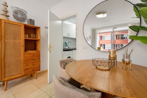 A dining area with a round wooden table, a wicker cabinet, and a mirror reflecting a building outside.