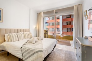 A hotel room with a bed, a desk, a chair, a balcony with a view of a building, and a window with curtains.