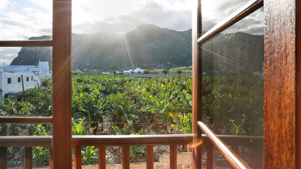 Blick durch ein Fenster mit Holzrahmen auf eine üppige Bananenplantage, entfernte Berge und ein weißes Gebäude.