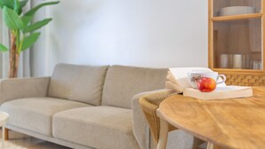 A living room with a sofa, a wooden table with a book and a fruit, and a bookshelf in the background.