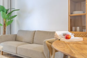 A living room with a sofa, a wooden table with a book and a fruit, and a bookshelf in the background.
