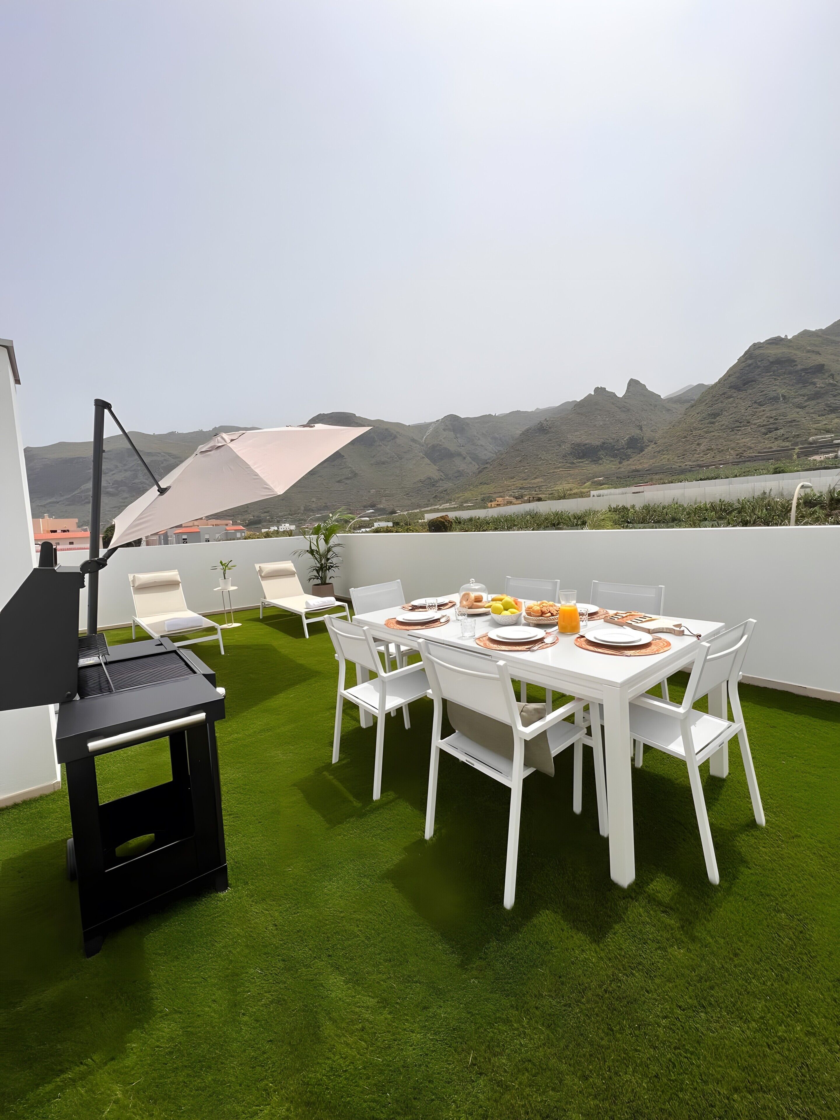 Terraza en la azotea con una mesa de comedor lista para cuatro personas, rodeada de sillas blancas, una parrilla y un paraguas grande.
