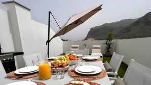 Ein Esstisch draußen mit Tellern, Besteck, einer Holzplatte mit Käse, Früchten und einem Glas Orangensaft.
