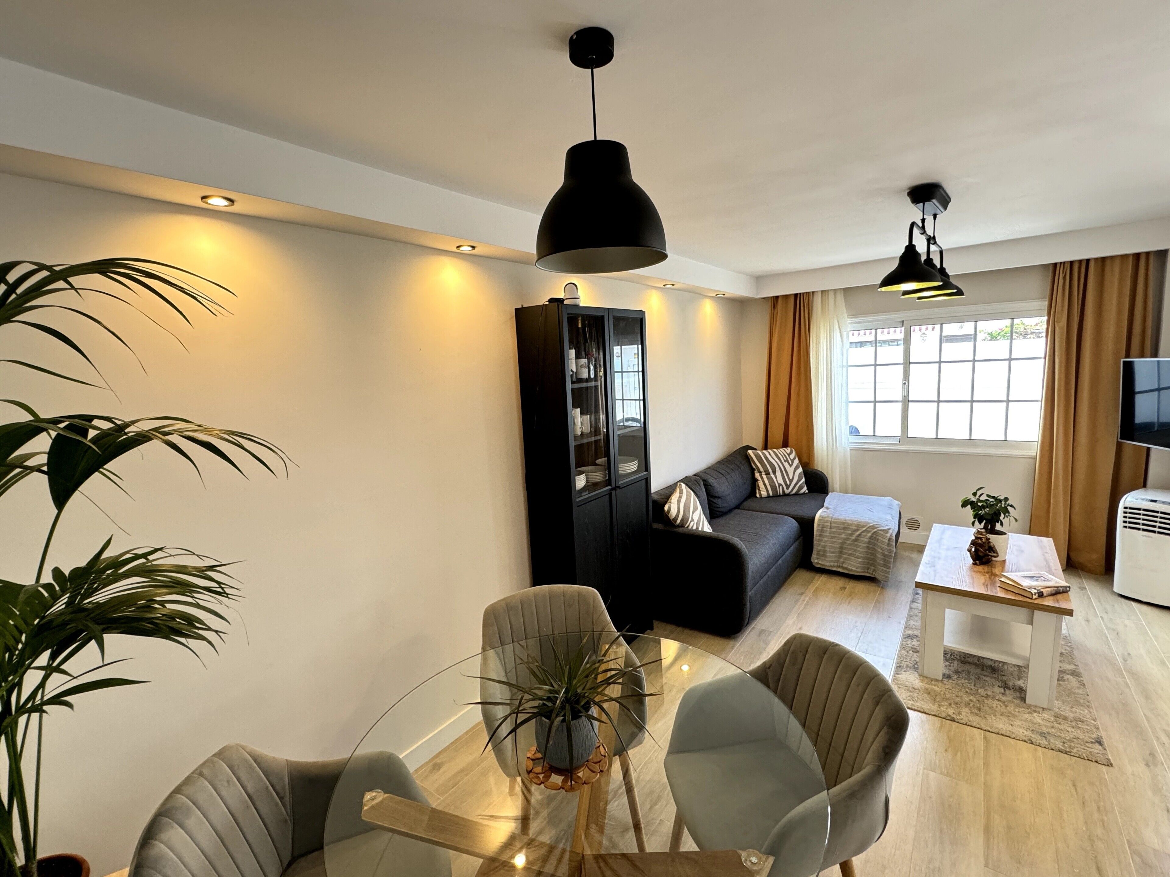 Uma sala de estar moderna com uma mesa de centro de vidro, poltronas cinzas, um sofá preto, um armário e uma TV.