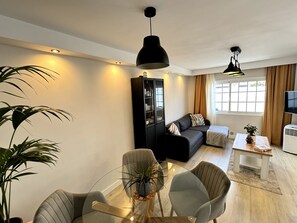Un salon moderne avec une table basse en verre, des fauteuils gris, un canapé noir, un meuble et un téléviseur.