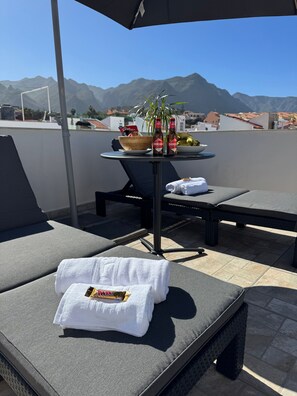 Une terrasse sur le toit, avec des chaises longues, une table avec des bouteilles, un saladier rempli de fruits et des serviettes.