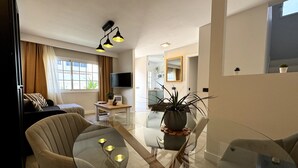 Ein modernes Wohnzimmer mit einem Glastisch, zwei Stühlen, einer Couch, einem Fernseher und einer Topfpflanze.
