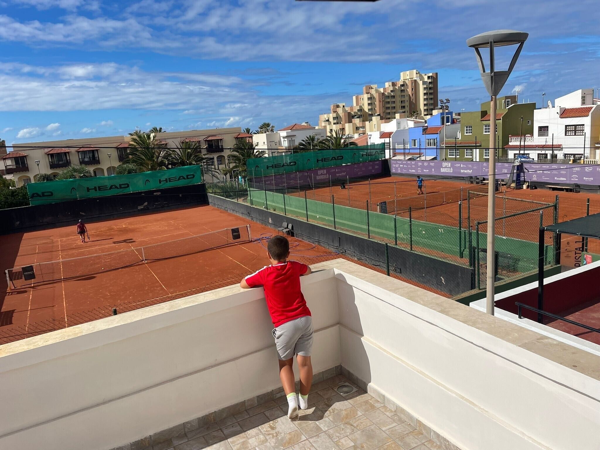 Una cancha de tenis con una persona apoyada en una pared, rodeada de edificios y un cielo despejado.