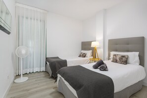 A hotel room with two beds, a standing fan, a lamp, and a window with sheer curtains.