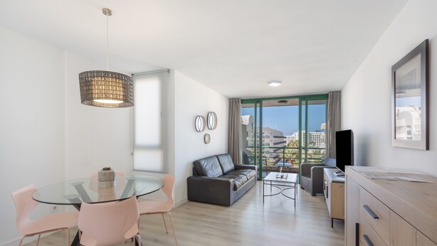 A modern living room with a glass dining table, a sofa, a TV, and a view of the city.