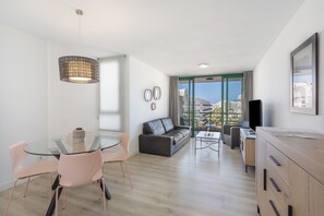 A modern living room with a glass dining table, a sofa, a TV, and a view of the city.
