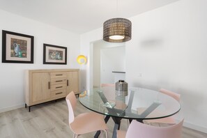 A modern dining area with a glass table, pink chairs, a wooden cabinet, and two framed artworks on the wall.
