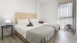 A hotel room with two beds, a bedside table with a lamp, a standing fan, and sheer curtains.
