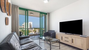 A modern living room with a leather sofa, a glass coffee table, a TV on a wooden cabinet, and a balcony with a view of buildings.