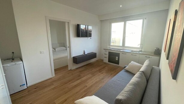 A modern, well-lit living area with a sofa, a small kitchenette, and a window providing natural light. - Flora: 1 Bedroom Apartment 1 (Milano)