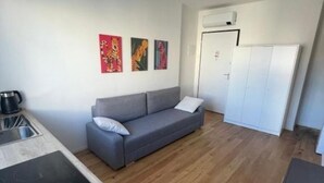 A small, modern room with a grey sofa, a white wardrobe, and a kitchenette with a stove. - Flora: 1 Bedroom Apartment 1 (Milano)