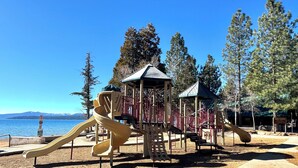 Un parc infantil amb tobogan i elements per escalar, envoltat d'arbres i amb vista a un llac.