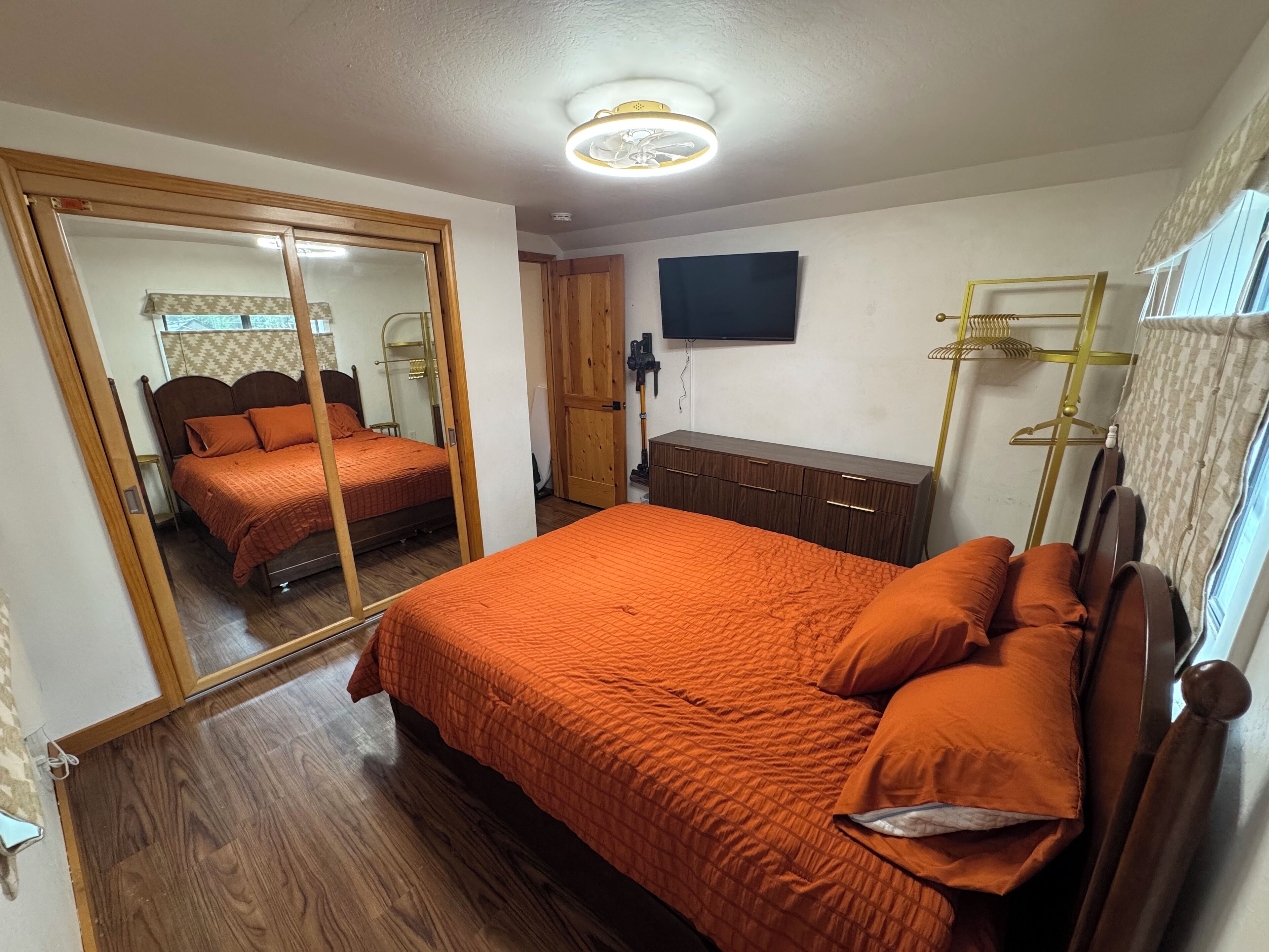 Un dormitorio con un espejo grande, una cama con ropa de cama naranja, un tocador de madera y un televisor en la pared.