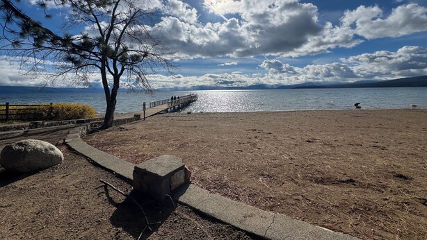 Una platja amb una passera de fusta, una banca de pedra i un arbre.