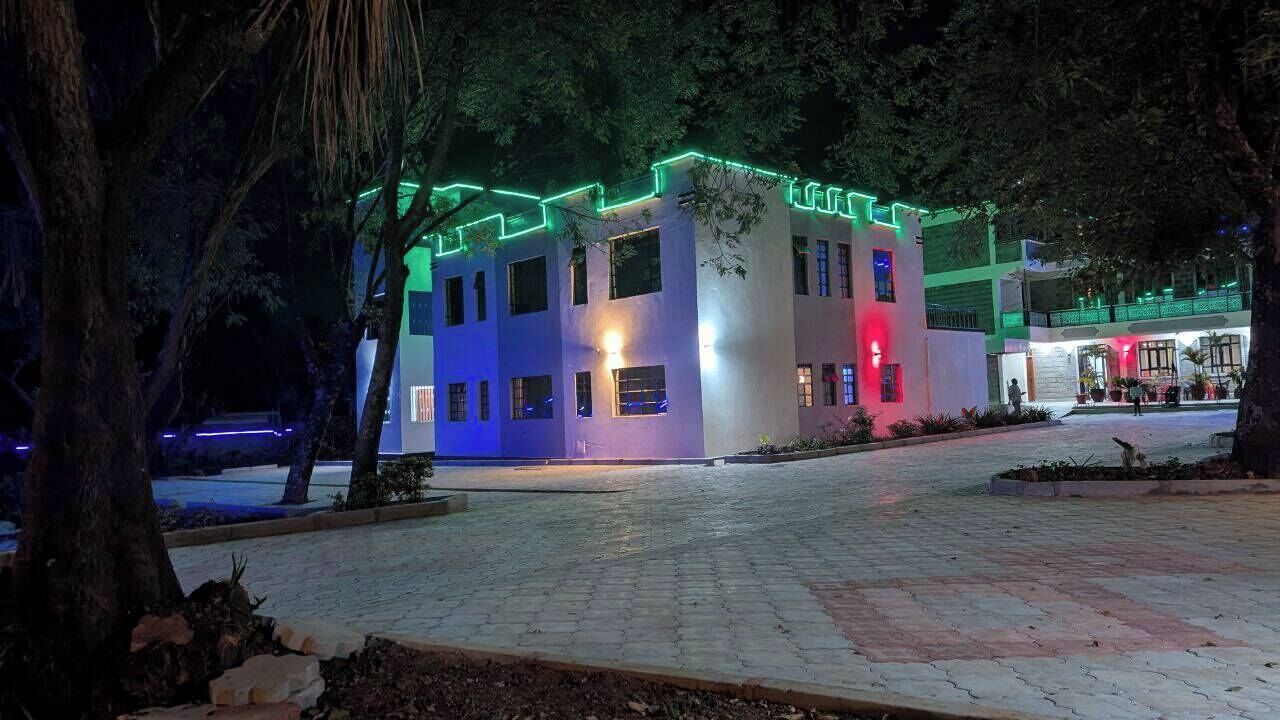 A multi-story building with illuminated green and blue lights, surrounded by trees and a paved area.