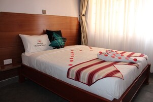 Habitación de hotel con cabecera de madera, cama con cubrecama blanco y detalles rojos, almohada con logotipo y ventana con cortinas.
