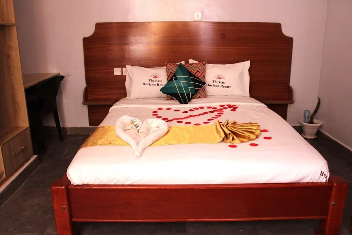 A neatly made bed with a heart-shaped towel arrangement, a golden throw blanket, and red rose petals on a white bedspread.