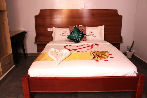 A neatly made bed with a heart-shaped towel arrangement, a golden throw blanket, and red rose petals on a white bedspread.