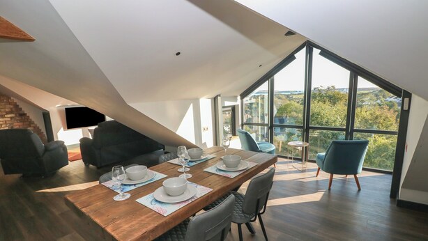 Un espace repas avec une table en bois dressée pour quatre personnes, entourée de chaises modernes, dans un loft spacieux doté de grandes fenêtres offrant une vue sur la verdure.