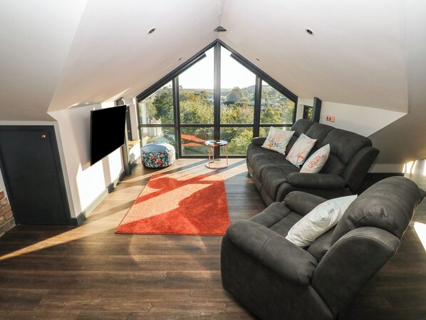 A modern loft bedroom with a large skylight, a dark leather sofa, a red rug, and a flat-screen TV mounted on the wall. - 11 Brython Place (Tenby)