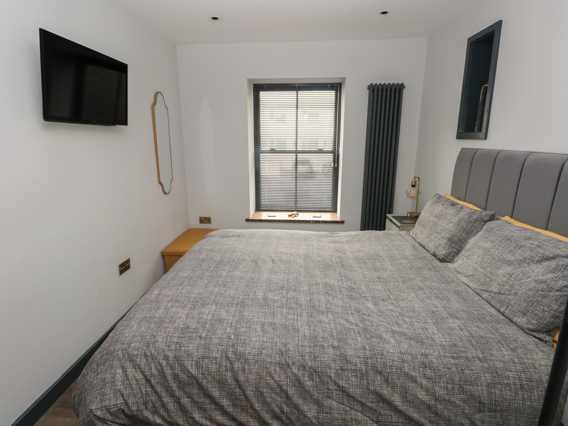 A bedroom with a bed, a window with blinds, a bedside table, and a mounted TV.
