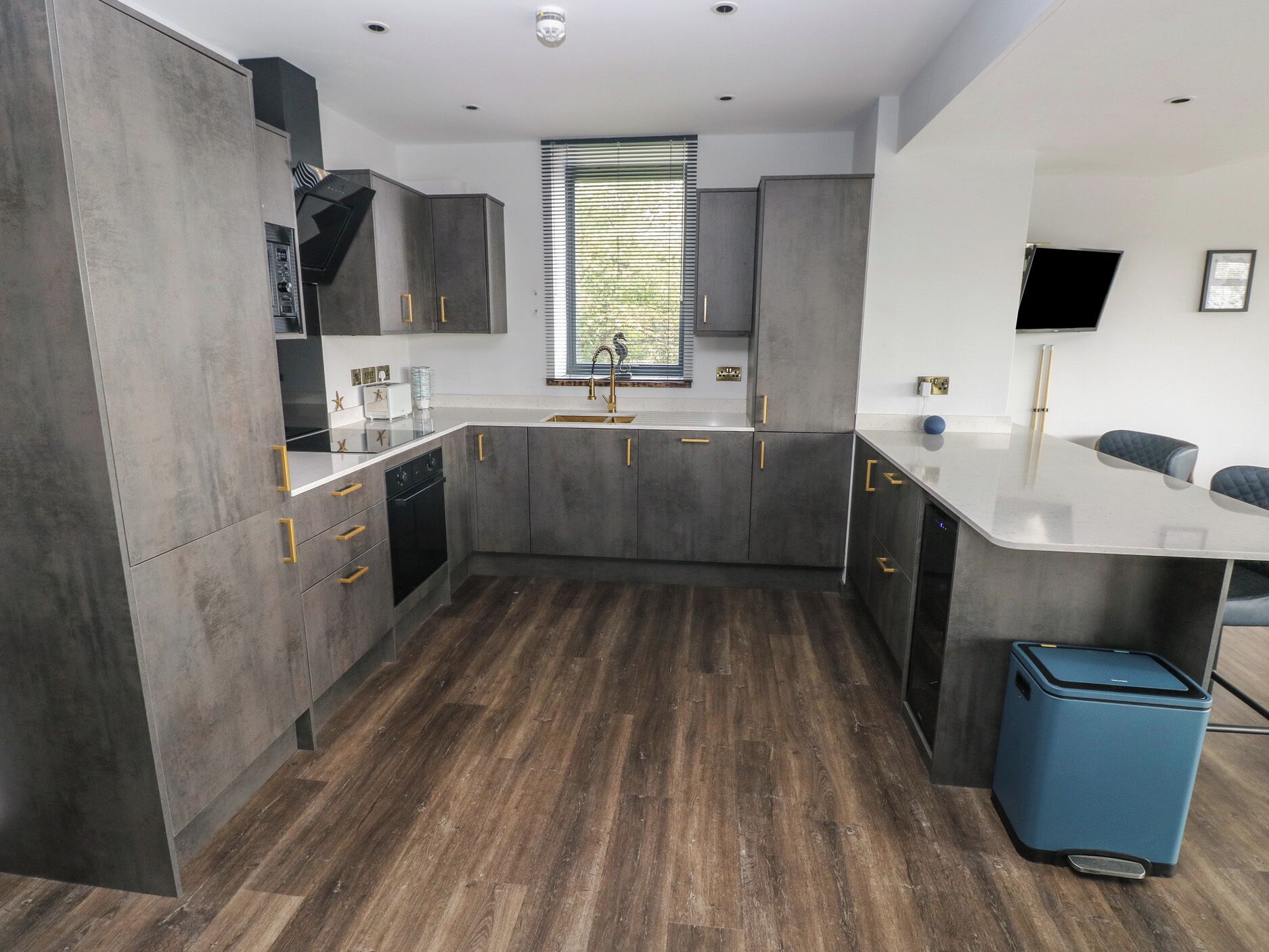 A modern kitchen with grey cabinets, a built-in oven, and a blue trash bin.