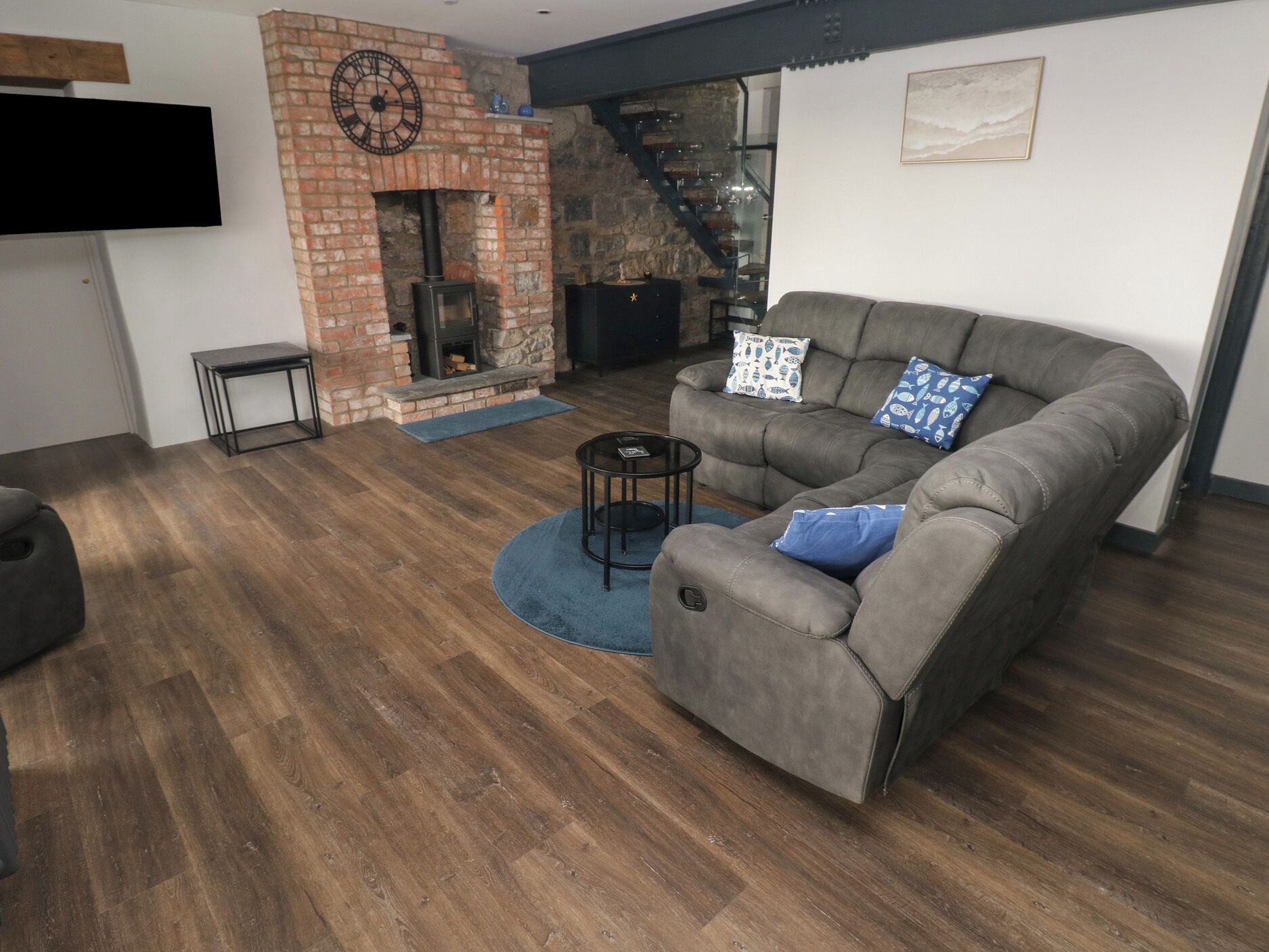 A modern living room with a brick fireplace, a grey sofa with patterned cushions, and a flat-screen TV mounted on the wall.