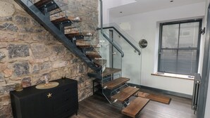 A modern staircase with glass railings and wooden steps, leading to a room with a stone wall and a black dresser with drawers. - 10 Brython Place (Tenby)