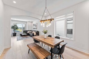 A modern dining area with a wooden table and benches, a hanging light fixture, and a view of the outdoors through a large window. - *NEW* Prestigious 5BED Home | 25MINS 2DT (Ottawa)