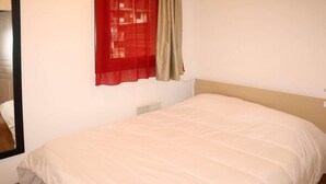 Un dormitorio con cama, una ventana con cortinas rojas y beige, y una pared blanca.