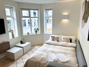 Un dormitorio con una cama grande, mesitas de noche, un televisor de pantalla plana y ventanas con vistas a la calle.