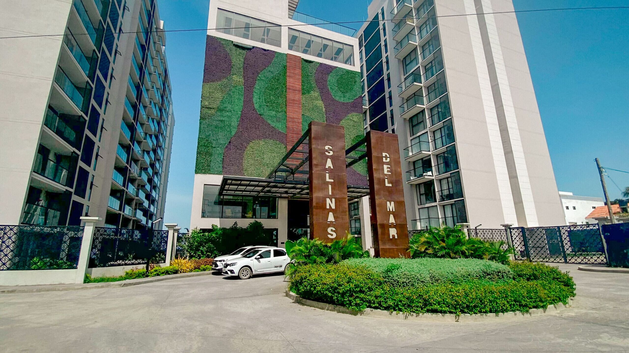 A modern building with a green patterned facade, two vertical signposts with 'SALINAS DEL MAR' text, and a white car parked in front.