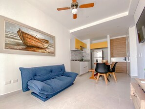 A modern living room with a blue sofa, a ceiling fan, a kitchen area with yellow cabinets, and a dining table with chairs.