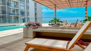 Uma área à beira da piscina com espreguiçadeiras, um vaso de concreto com flores e vista para um edifício moderno.