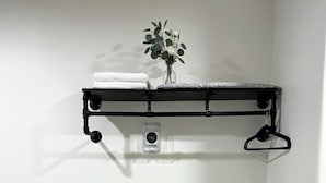 Una sala moderna con un sofá gris, un estante de metal negro con un jarrón y toallas, y un cubo de basura negro pequeño.