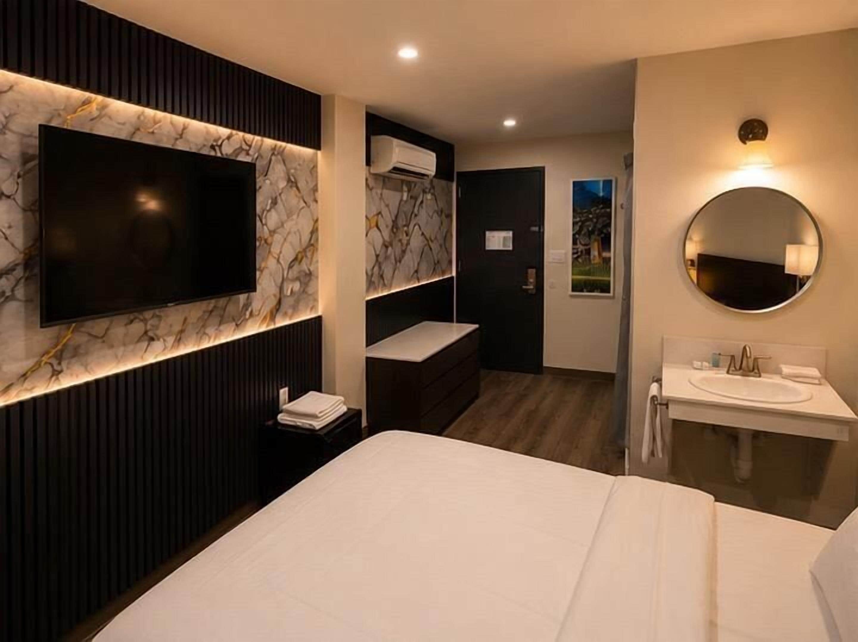 A modern hotel room with a large bed, a TV mounted on a marble wall, a round mirror above the sink, and a view of the outdoors through a window.