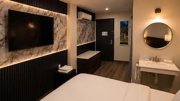 A modern hotel room with a large bed, a TV mounted on a marble wall, a round mirror above the sink, and a view of the outdoors through a window.