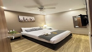 Una habitación de hotel moderna con una cama grande, un ventilador de techo, un cuadro de un paisaje urbano, una mesita de noche, una lámpara y un espejo.