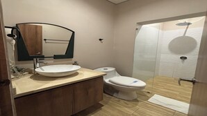 Un baño moderno con tocador de madera, un lavamanos blanco, inodoro y cabina de ducha con mampara de vidrio.
