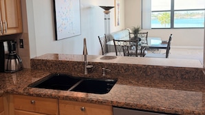 A kitchen with granite countertops, a double sink, wooden cabinets, and a stainless steel dishwasher. There is a dining area with a table and chairs, and a large window offering a view of the ocean. - Charming condo in fantastic St. Petersburg Beach, with waterfront, sunset views (St. Petersburg)